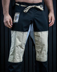 A man wearing a pair of martial arts pants with a black and beige color-blocked design, featuring a cream-colored drawstring and a brand patch on the left thigh, standing barefoot against a dark paneled background.