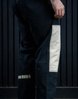 A person shown from the waist down wearing loose black martial arts pants with a large off-white vertical patch on the right leg and a small gold BUD logo near the lower left leg, standing barefoot against a dark paneled wall.