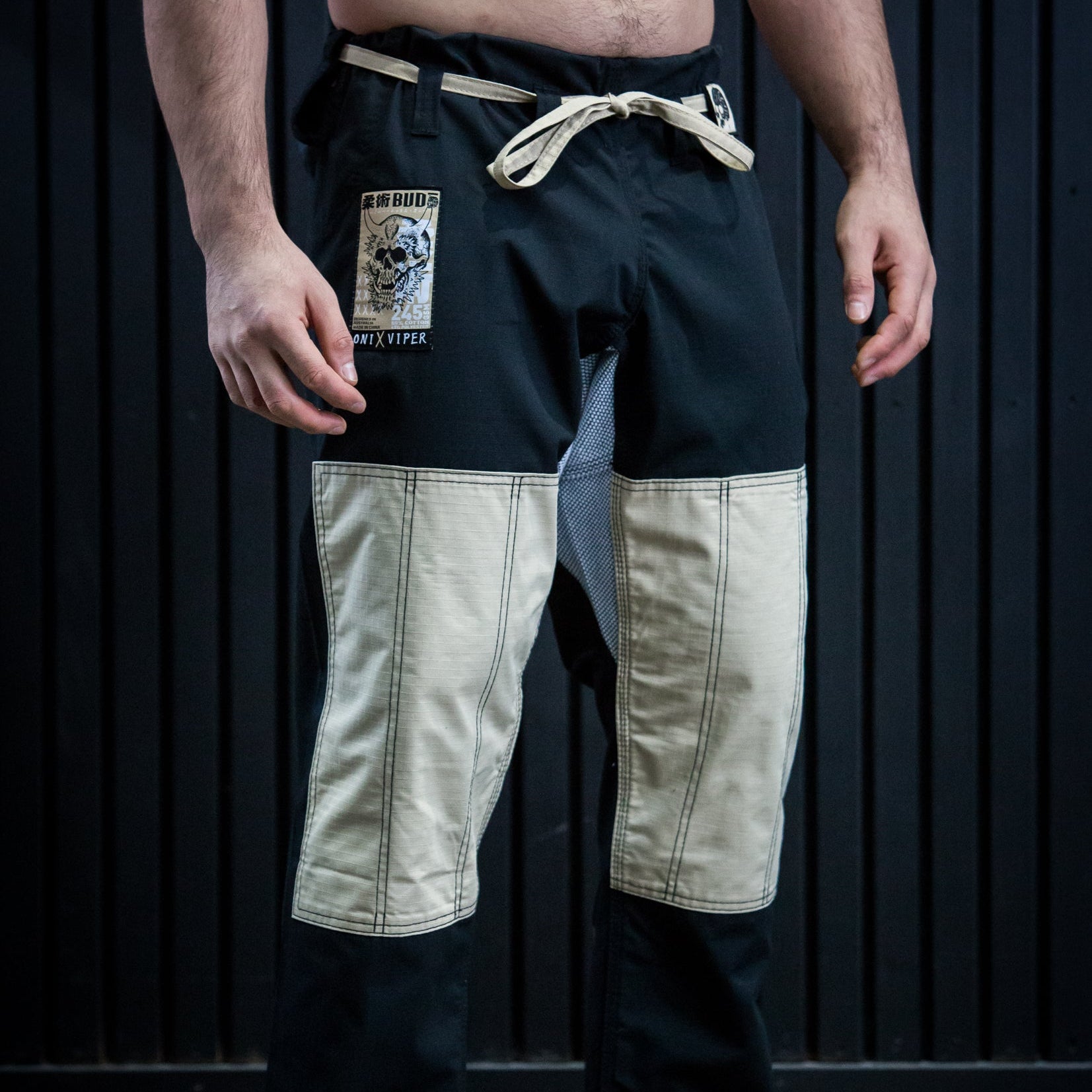 A man wearing black martial arts pants with beige patchwork leg panels and a cream-colored drawstring, standing barefoot against a dark paneled background.