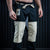 A man wearing black martial arts pants with beige patchwork leg panels and a cream-colored drawstring, standing barefoot against a dark paneled background.
