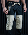 A man wearing black martial arts pants with beige patchwork leg panels and a cream-colored drawstring, standing barefoot against a dark paneled background.