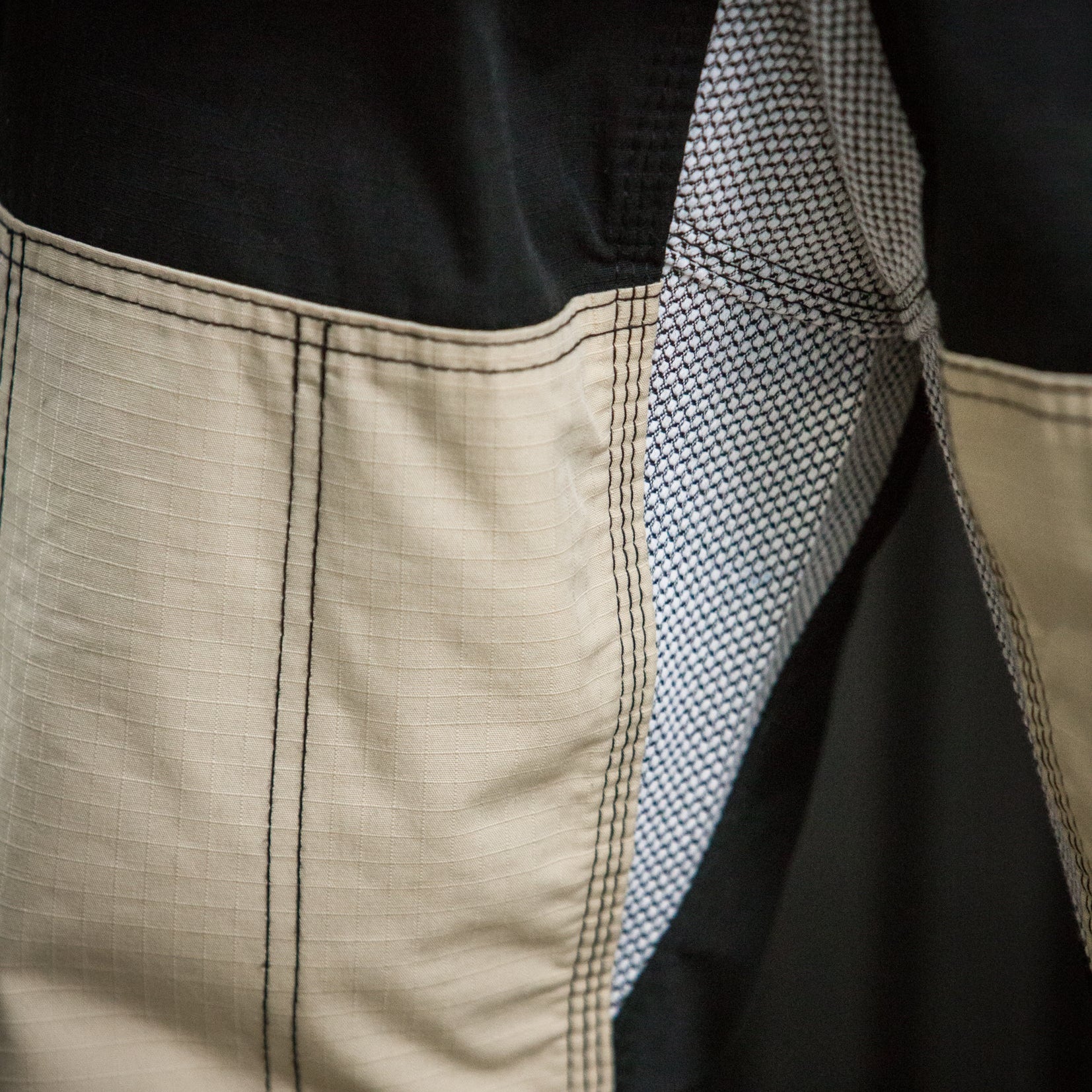 Close-up detail of a garment showing a beige fabric panel with black double-stitched seams, a black interior lining, and a textured gray-and-white mesh inner layer.