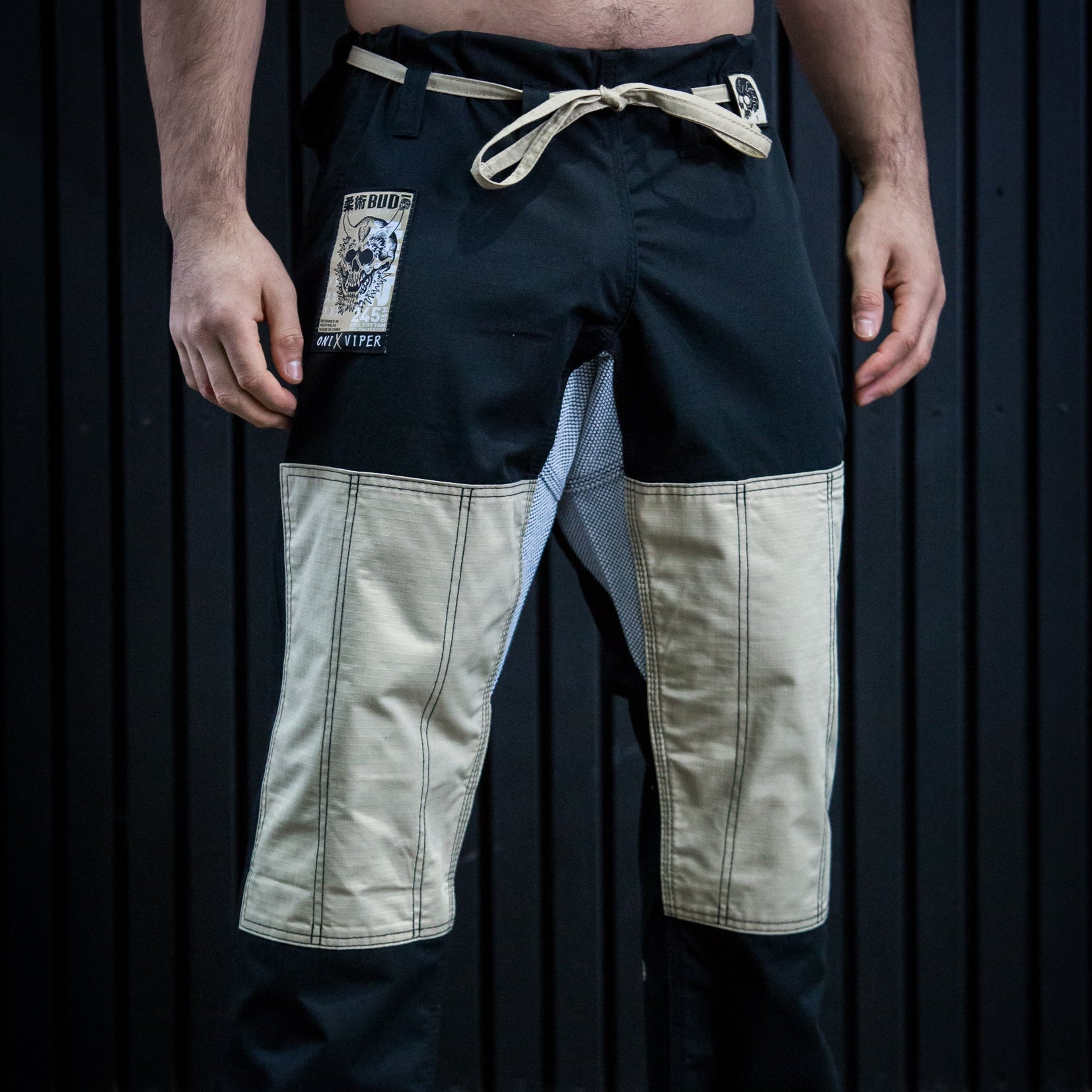A man wearing a pair of martial arts pants with a black and beige color-blocked design, featuring a cream-colored drawstring and a brand patch on the left thigh, standing barefoot against a dark paneled background.