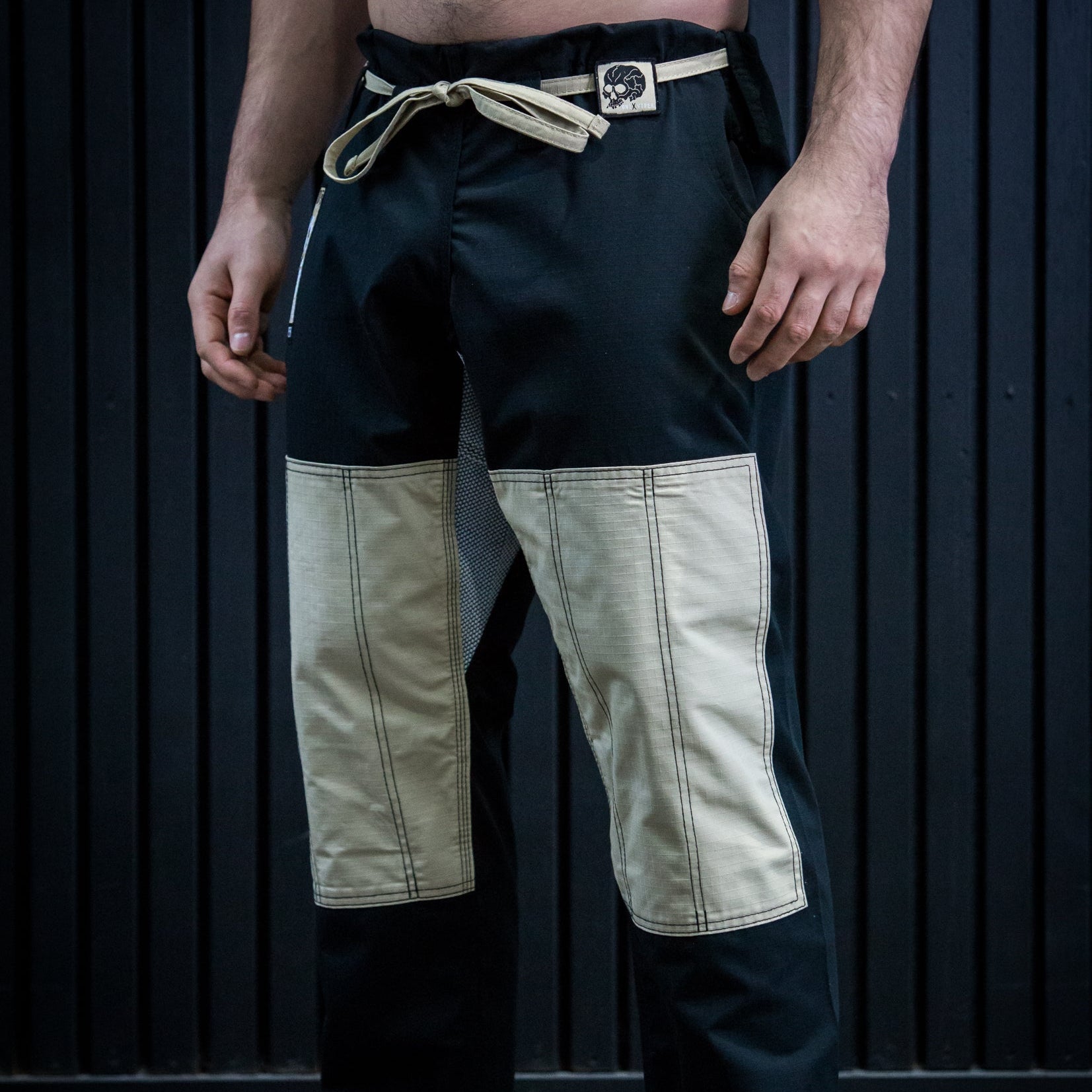 A man wearing black martial arts pants with beige color-blocked knee panels and a beige drawstring belt, standing barefoot against a dark paneled wall.