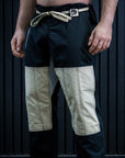 A man wearing black martial arts pants with beige color-blocked knee panels and a beige drawstring belt, standing barefoot against a dark paneled wall.