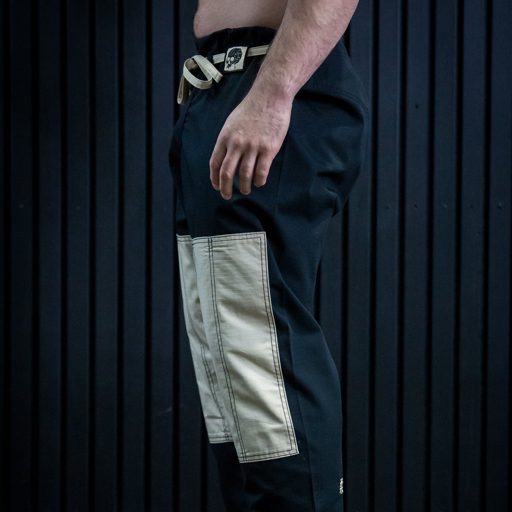 A side profile of a person wearing black martial arts pants with large beige patch pockets and a black belt, standing barefoot against a dark paneled wall.