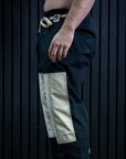 A side profile of a person wearing black martial arts pants with large beige patch pockets and a black belt, standing barefoot against a dark paneled wall.