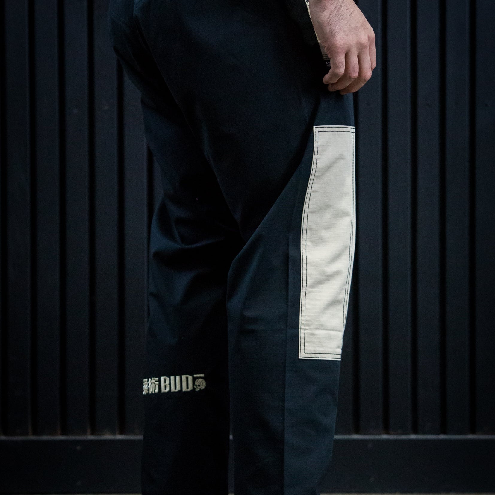 A person shown from the waist down wearing loose black martial arts pants with a large off-white vertical patch on the right leg and a small gold BUD logo near the lower left leg, standing barefoot against a dark paneled wall.